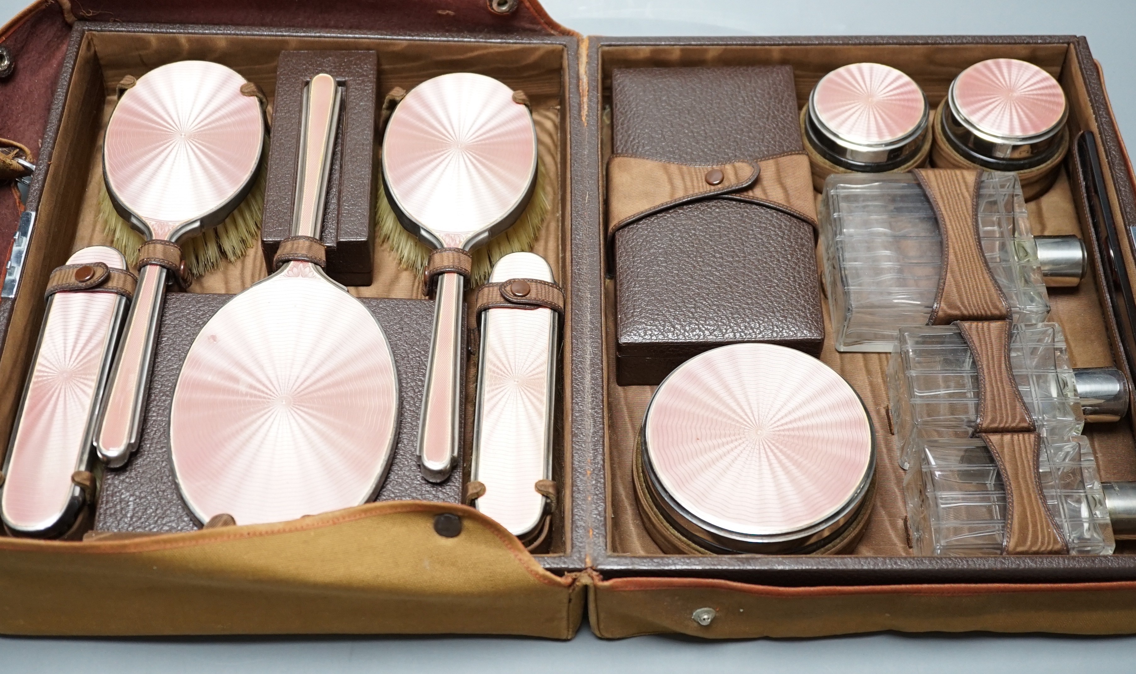 A George V travelling toilet case, containing eleven silver and pink enamel mounted glass bottles jars, mirror and brushes, Mappin & Webb, 1936, in brown leather case, case width 31cm.